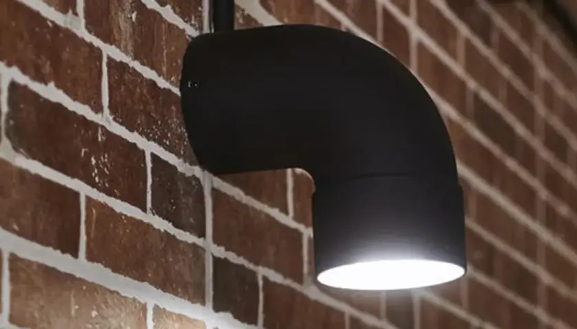 Applique fai da te in metallo nero a forma di gomito, illumina una parete in mattoni rossi.