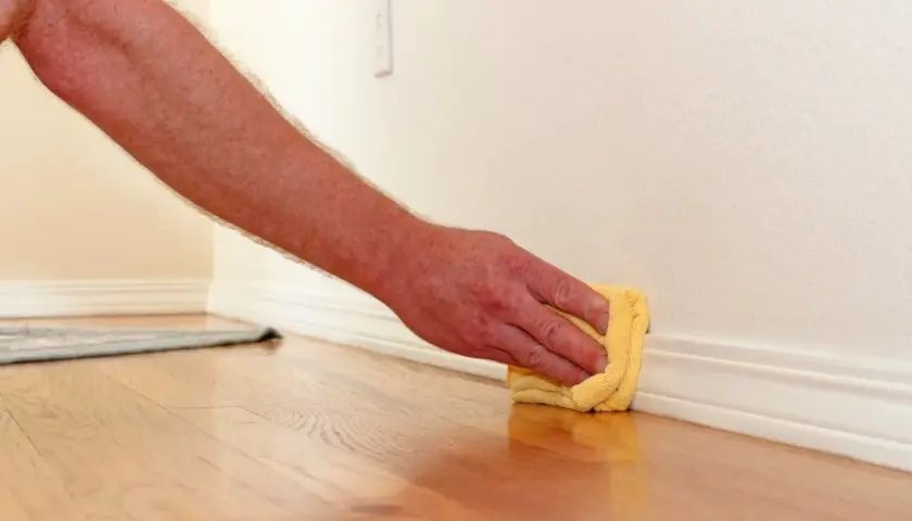 Mano limpiando rodapiés con un paño amarillo. El suelo de madera brilla y la pared está pintada de blanco.
