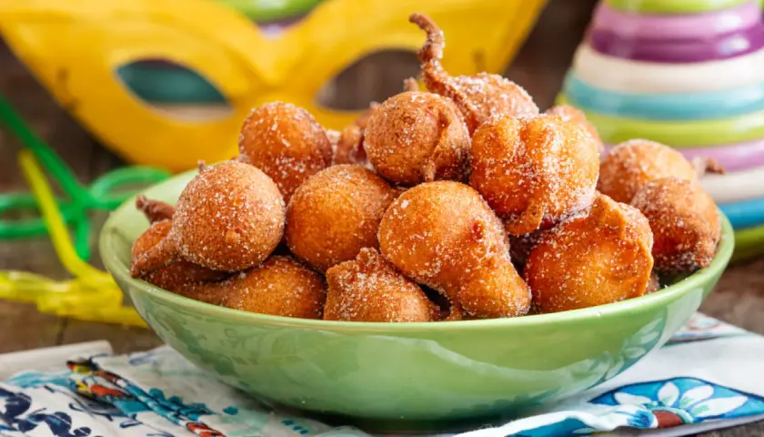 Frittelle di una volta, dolcetti dorati cosparsi di zucchero, pronti per il Carnevale. Maschere e coriandoli sullo sfondo.