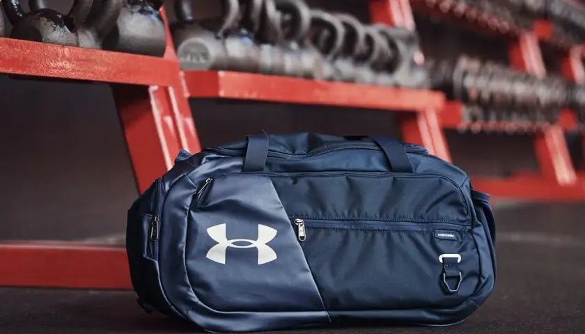 Granatowa torba sportowa 100l z logo Under Armour, gotowa na trening. W tle sztangi i hantle.