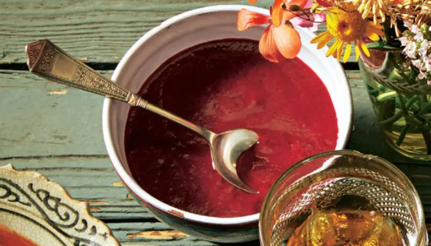 Ein Schälchen mit roter Soße, perfekt für ein Tkemali Rezept. Ein Löffel ruht darin, daneben ein Glas mit Bernstein-farbener Flüssigkeit und Blumen.