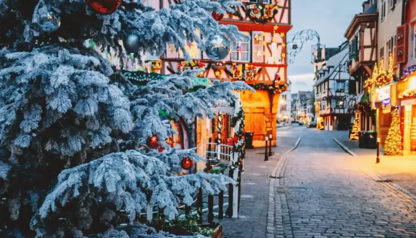 Verschneiter Weihnachtsbaum mit roten Kugeln vor Fachwerkhäusern, die mit Lichtern geschmückt sind. Perfekte reiseziele winter.