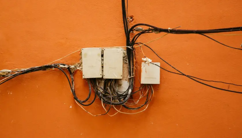 Stare skrzynki elektryczne z plątaniną kabli na pomarańczowej ścianie.