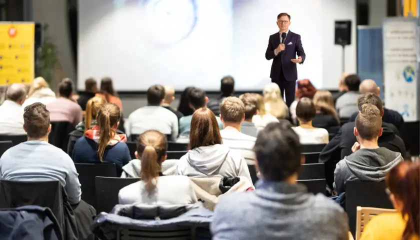 Mężczyzna w garniturze wygłasza przemówienie do publiczności siedzącej na sali.