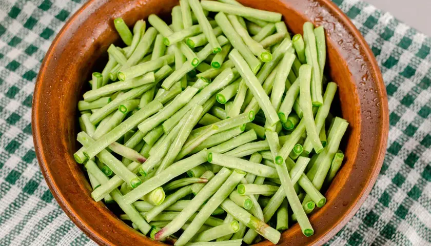 Fagiolini in umido freschi e verdi, pronti per essere cucinati in una ciotola di terracotta su una tovaglia a quadretti.