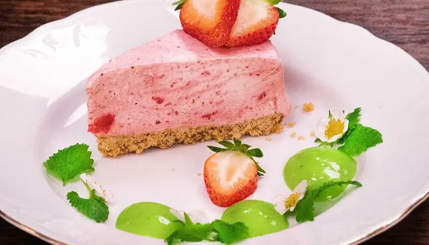 En bit krämig cheesecake jordgubb med färska bär och mynta på en vit tallrik.