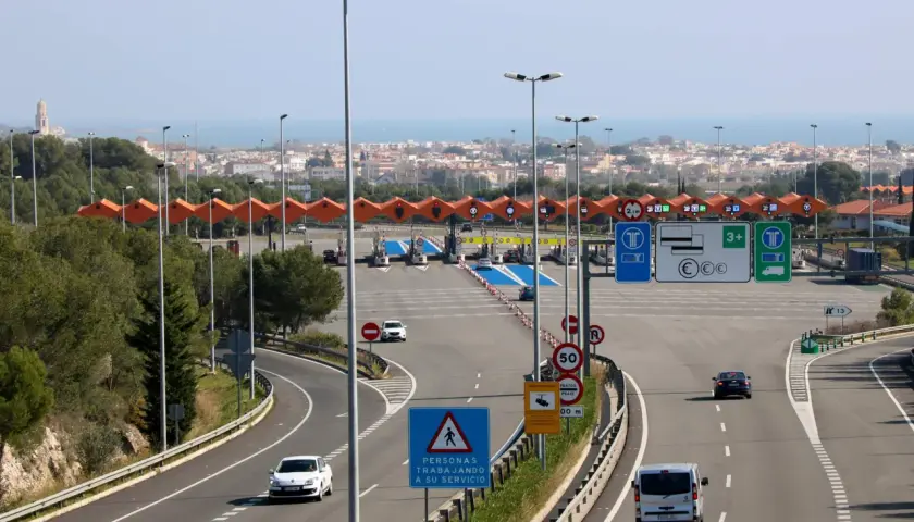 Peaje en la autopista Pau Casals con varios carriles, señales de tráfico y vehículos circulando. Al fondo, un paisaje urbano y el mar.