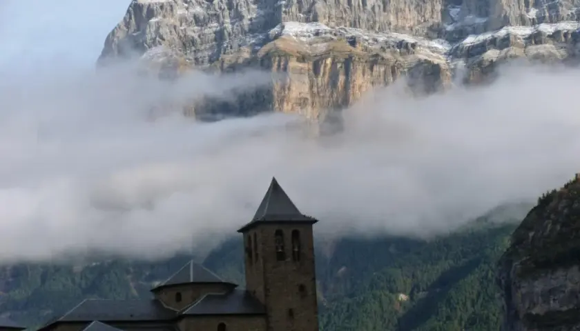 Iglesia románica con campanario, rodeada de niebla, bajo imponentes montañas nevadas. Ideal para explorar rutas desde Torla.