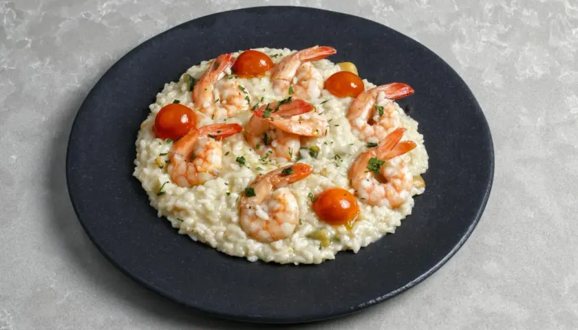 Kremowe risotto z warzywami, ozdobione krewetkami i pomidorkami koktajlowymi, posypane natką pietruszki.