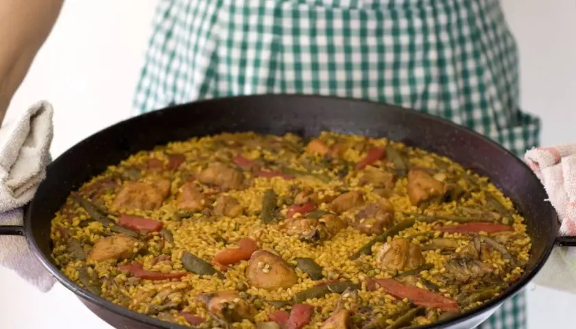 Una persona con un delantal a cuadros verdes sostiene una paella valenciana recién hecha, llena de arroz, pollo, judías verdes y pimientos rojos.