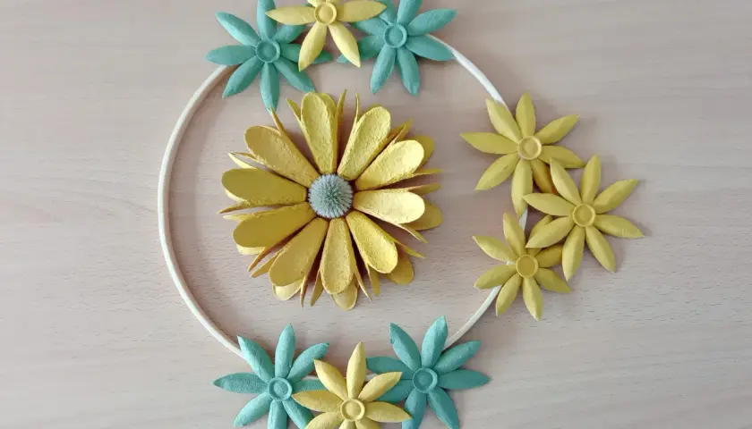 Kreative Blumen aus Eierkarton, gelb und türkis, um einen weißen Ring arrangiert.