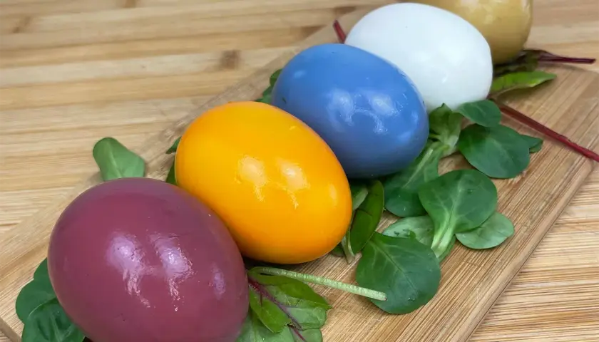 Cinq œufs colorés, du violet à l'orange vif, en passant par le bleu, le blanc et le doré, disposés sur un lit de verdure sur une planche en bois. Chaque couleur oeuf est une surprise.