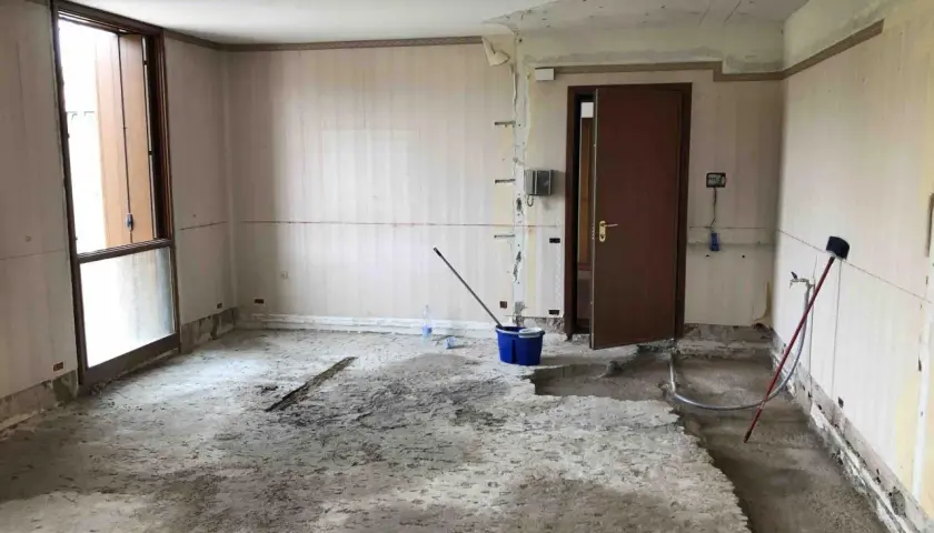 Interno di una stanza in ristrutturazione, con il pavimento in cemento grezzo e pareti spoglie. Si notano attrezzi da lavoro, tra cui un secchio blu e una scopa, che suggeriscono i lavori in corso e i possibili svantaggi dell'intonaco armato.