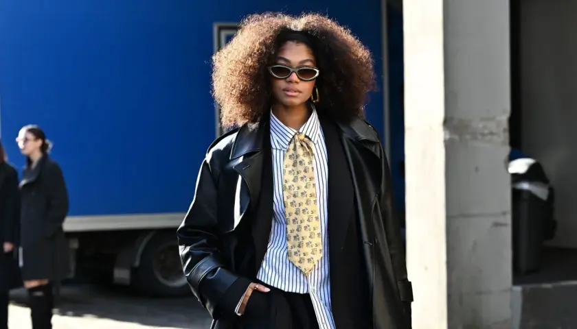 Une femme porte un pantalon noir, une chemise à rayures, une cravate fantaisie et un long manteau en cuir. Ses bottines sont cachées sous son pantalon.
