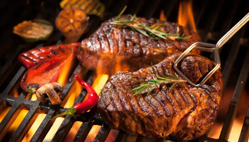 Deux steaks barbecue juteux, ornés de romarin, grésillent sur un barbecue avec des poivrons rouges, des champignons et des piments.