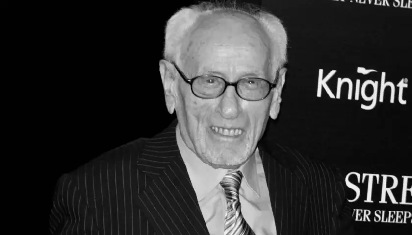 Eli Wallach, w okularach i garniturze w prążki, uśmiecha się. Ikona kina, pamiętana z niezapomnianych ról w filmach.