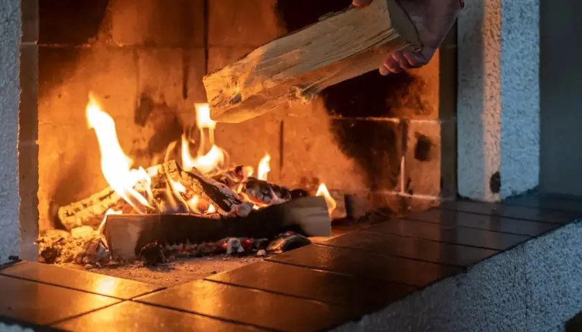 Eine Hand legt Holz nach, das im Kaminfeuer brennt. Das Holz brennt nicht, es spendet Wärme.