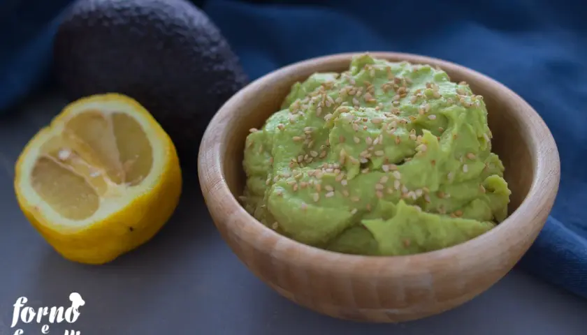 Hummus di avocado cremoso in ciotola di legno, guarnito con semi di sesamo. Accanto, un avocado intero e mezzo limone.