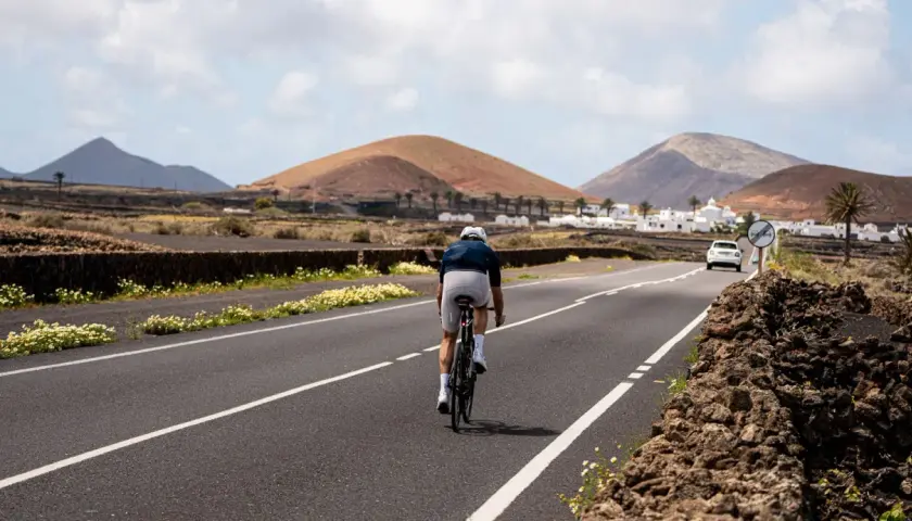 Ciclista pedaleando por una carretera en Lanzarote, con volcanes y casas blancas al fondo. Una aventura de lanzarote en bici.