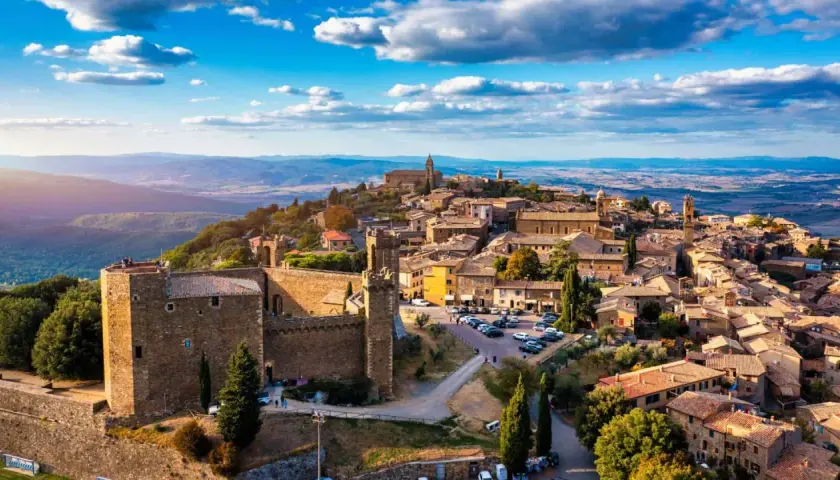 A panoramic view of medieval towns in Italy, perched on a hilltop under a bright blue sky with fluffy clouds.