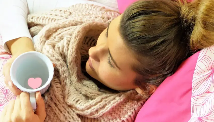 Donna avvolta in una sciarpa calda, tiene una tazza con un cuore rosa. Forse sente freddo in gravidanza, maschio o femmina, cerca conforto.