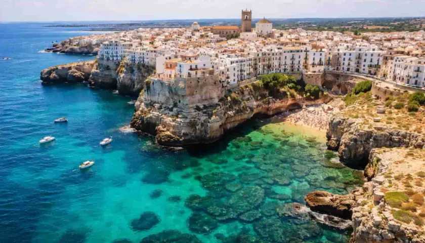 Vue aérienne de Polignano a Mare, avec ses maisons blanches perchées sur des falaises rocheuses surplombant une mer turquoise et une plage bondée.