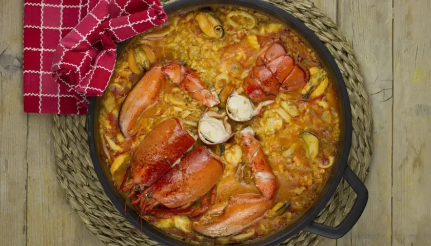 Paella de marisco con langosta, mejillones y calamares, servida en una paellera sobre una esterilla de mimbre.