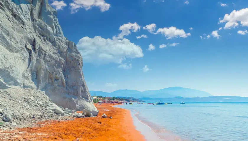 Ein atemberaubender Kefalonia Strand mit leuchtend orangefarbenem Sand, der von einer steilen weißen Klippe eingerahmt wird. Das türkisfarbene Meer lädt zum Schwimmen ein.