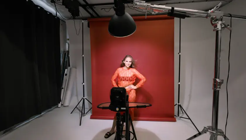 Modelka w pomarańczowej sukience pozuje na tle czerwonego tła. To profesjonalna fotografia studyjna z oświetleniem i sprzętem.