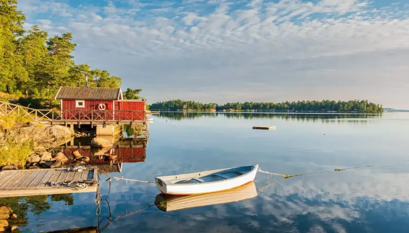 En vit båt ligger förtöjd vid en röd sjöbod. Kanske är det en båt till Drottningholm, redo för en lugn tur på vattnet.