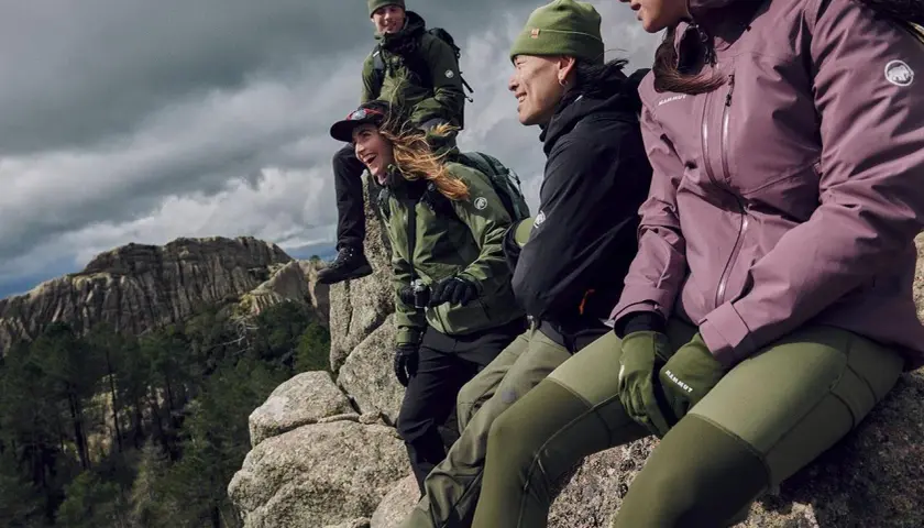 Grupa przyjaciół w zielonych i fioletowych kurtkach softshell siedzi na skale, podziwiając górski krajobraz.