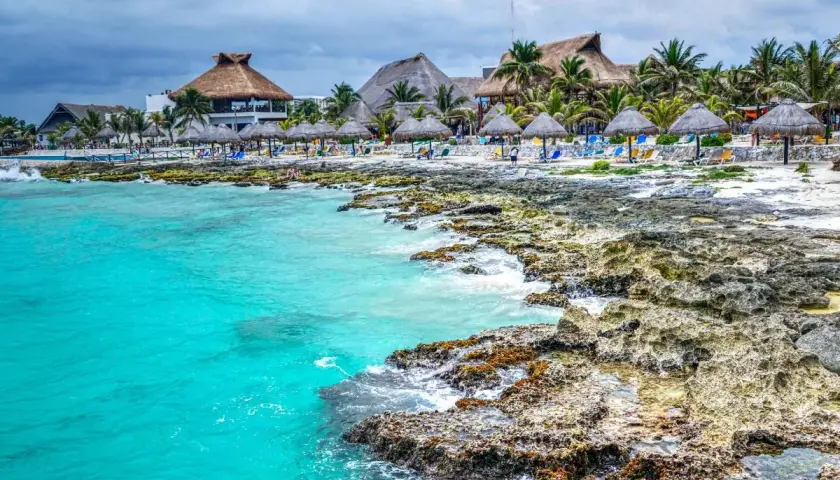 Plage mexicaine avec eau turquoise, rochers, palmiers et bungalows à toit de chaume.