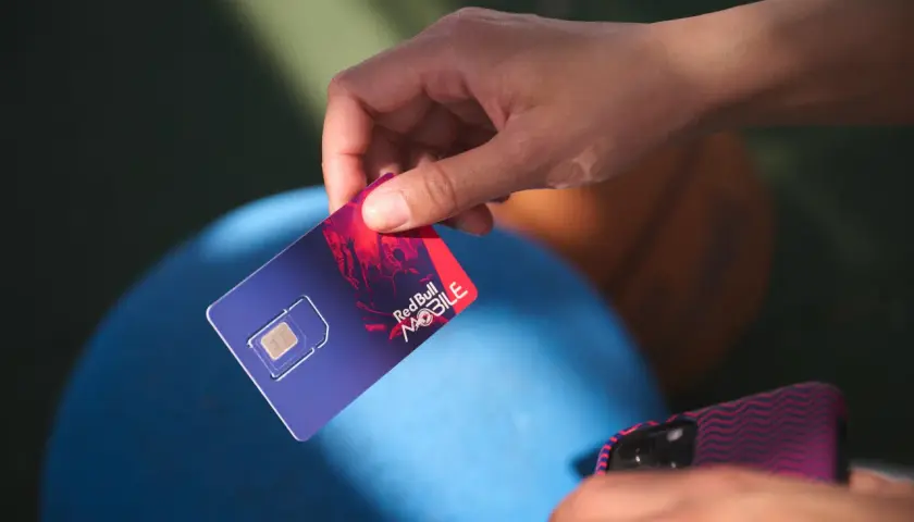 Dłoń trzyma kartę SIM Red Bull MOBILE, gotową do podróży po Europie. Internet na wyciągnięcie ręki!