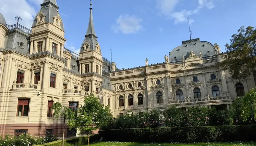 Imponujący pałac poznańskiego, z wieżyczkami i bogatymi zdobieniami, otoczony zielonym ogrodem z kwitnącymi krzewami róż.