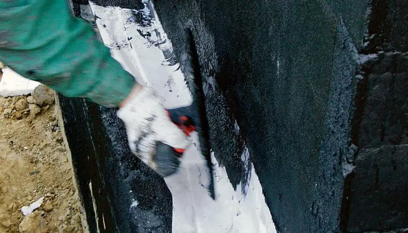 Ręka w rękawicy nakłada białą masę na czarną, wilgotną ścianę. To szlam mineralny, który chroni przed wodą.