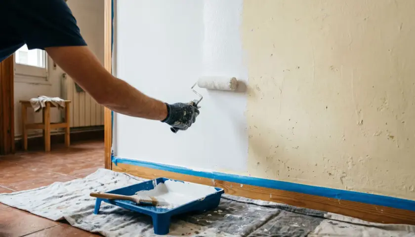 Persona con guantes pinta pared de blanco con rodillo. Bandeja con pintura y brocha lista para seguir como pintar una pared.