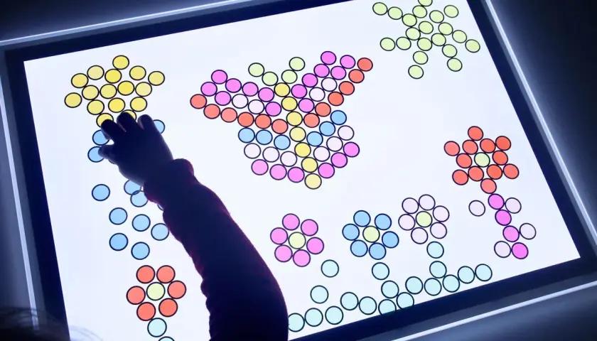 Niño/a creando figuras con piezas de colores sobre una mesa de luz, fomentando sus actividades creativas.