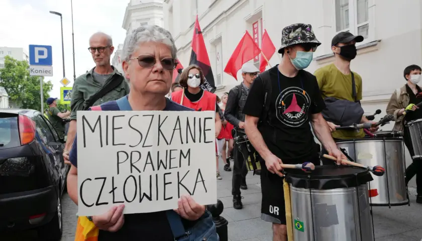 Kobieta trzyma transparent: "MIESZKANIE PRAWEM CZŁOWIEKA". W tle protestujący z bębnami i flagami.
