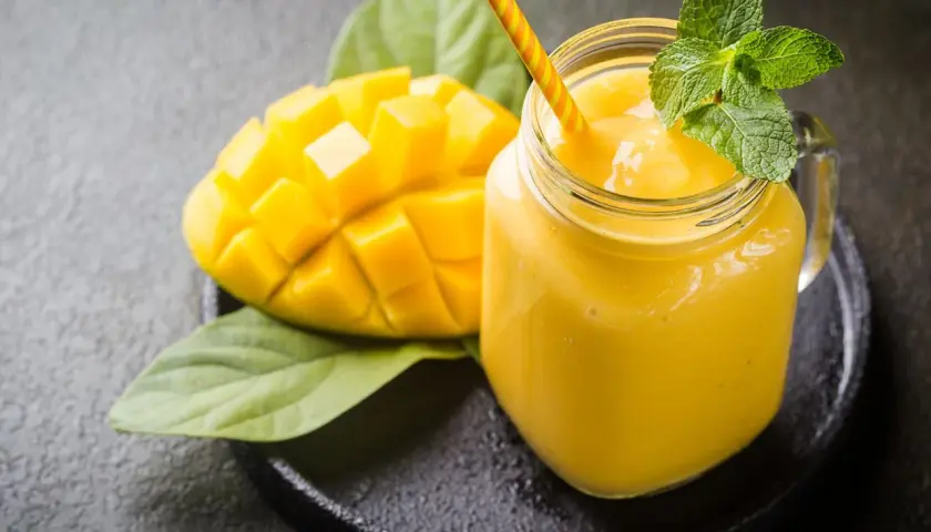 Un smoothie mangue onctueux dans un pot en verre, garni de menthe fraîche, à côté d'une moitié de mangue coupée en dés.