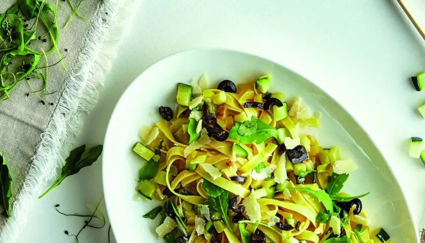 Pasta con verduras frescas, aceitunas negras y queso rallado, servida en un plato blanco.