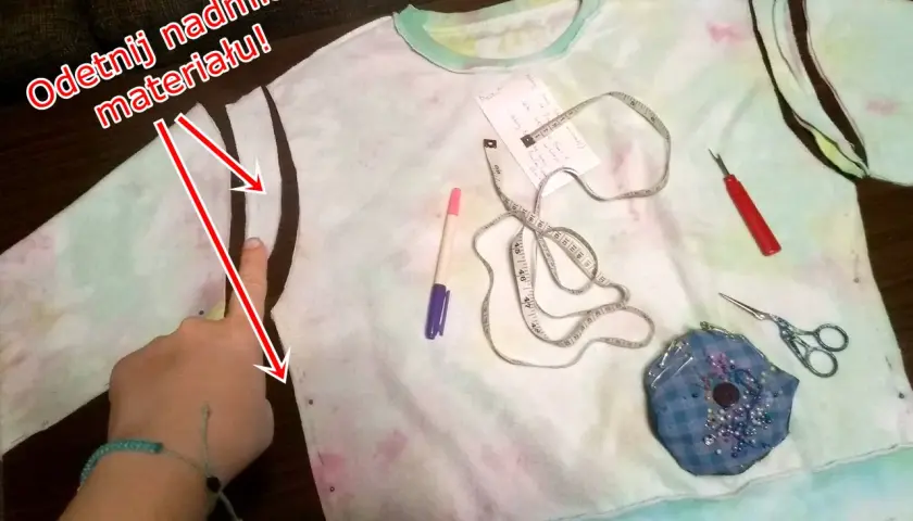 Ręka wskazuje odcięty rękaw bluzy, obok miarka, nożyczki i pisak.