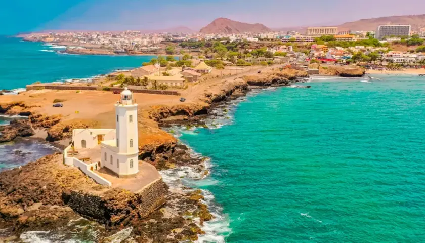 Faro blanco en una costa rocosa con aguas turquesas. Un pueblo costero se extiende detrás. ¡Ideas de qué hacer en Cabo Verde!