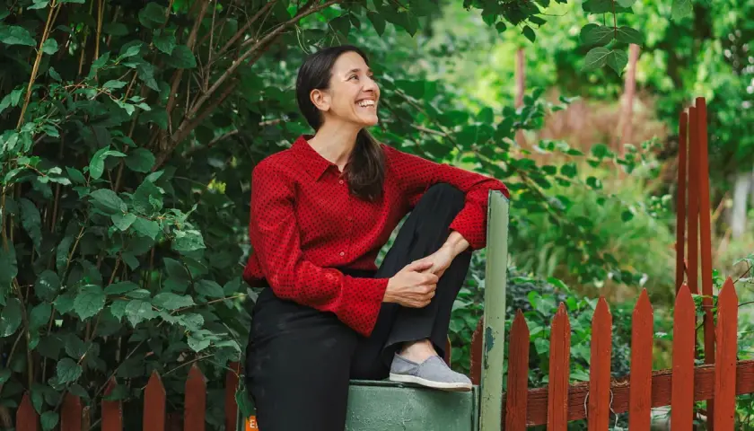 Paloma Grandin sitter avslappnat vid ett staket, omgiven av grönska. Hon ler och tittar uppåt, fångad i ett ögonblick av glädje.