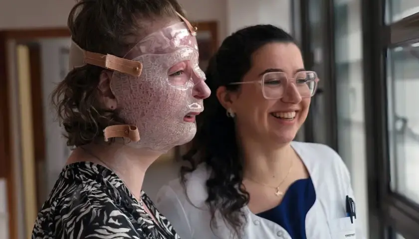 Une femme porte un masque transparent sur le visage, peut-être pour aider à comment cacher une brûlure. Une autre femme, souriante, la regarde.