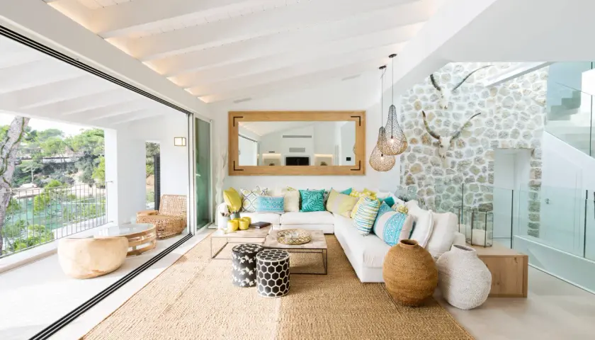 Helles Wohnzimmer mit Finca Style Einrichtung, weißem Sofa, bunten Kissen, Jute-Teppich und Blick auf das Meer.