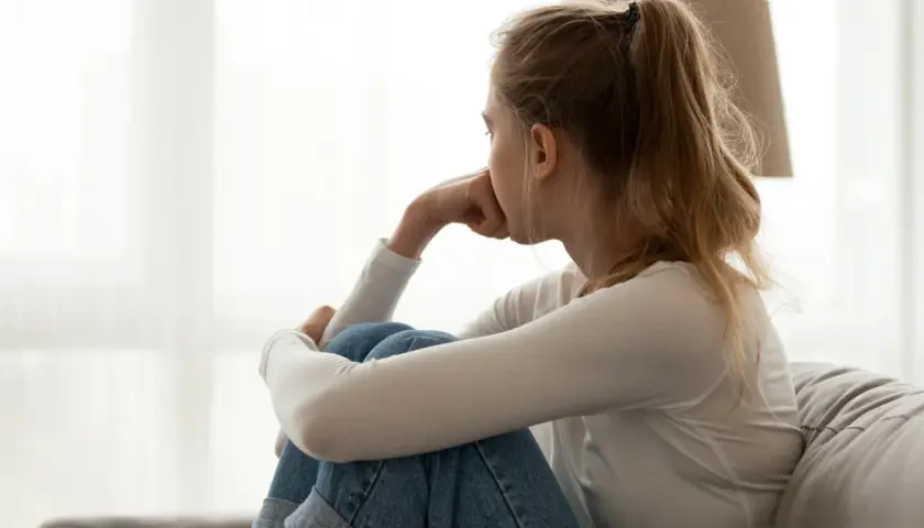 Młoda kobieta siedzi skulona na kanapie, zamyślona. Jej postawa sugeruje **tłumienie emocji, skutki** mogą być widoczne w jej wyrazie twarzy.