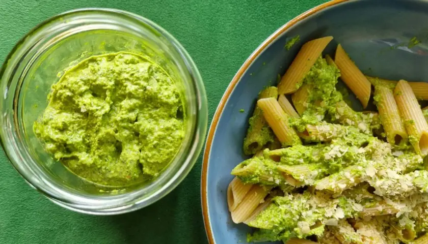 Pesto z liści rzodkiewki w słoiku obok talerza penne z tym zielonym sosem i startym parmezanem.