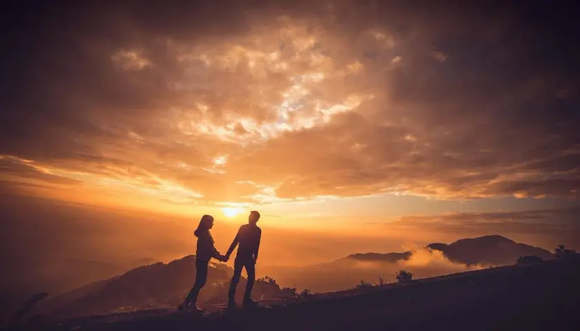 Para, trzymając się za ręce, podziwia zachód słońca na szczycie góry. Idealny romantyczny wypad we dwoje.
