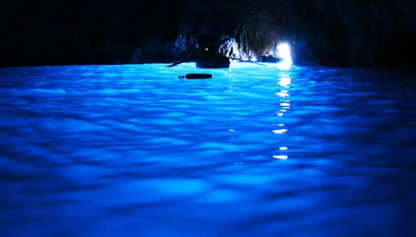Acqua blu elettrico nella grotta, un piccolo remo galleggia mentre una barca si dirige verso la luce all'uscita della blaue grotte.