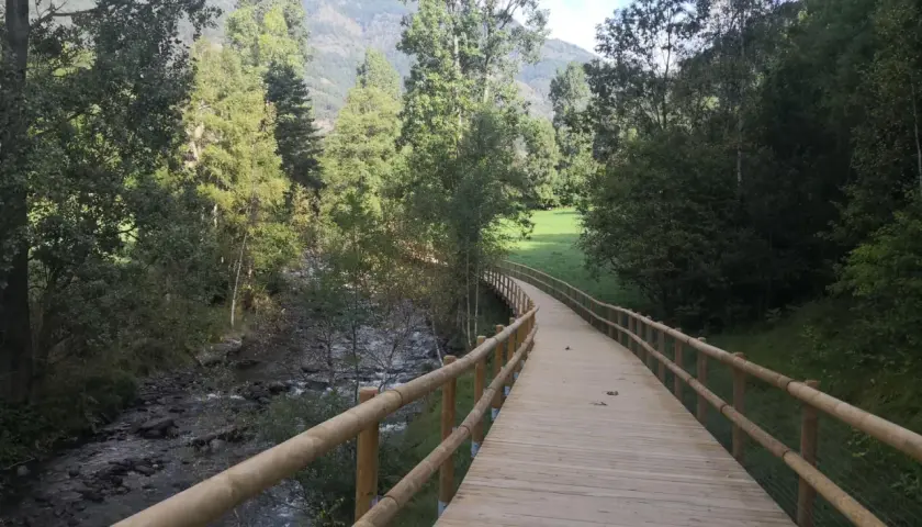 Paseo de madera junto a un río, parte de la ruta del ferro andorra, rodeado de árboles y montañas.